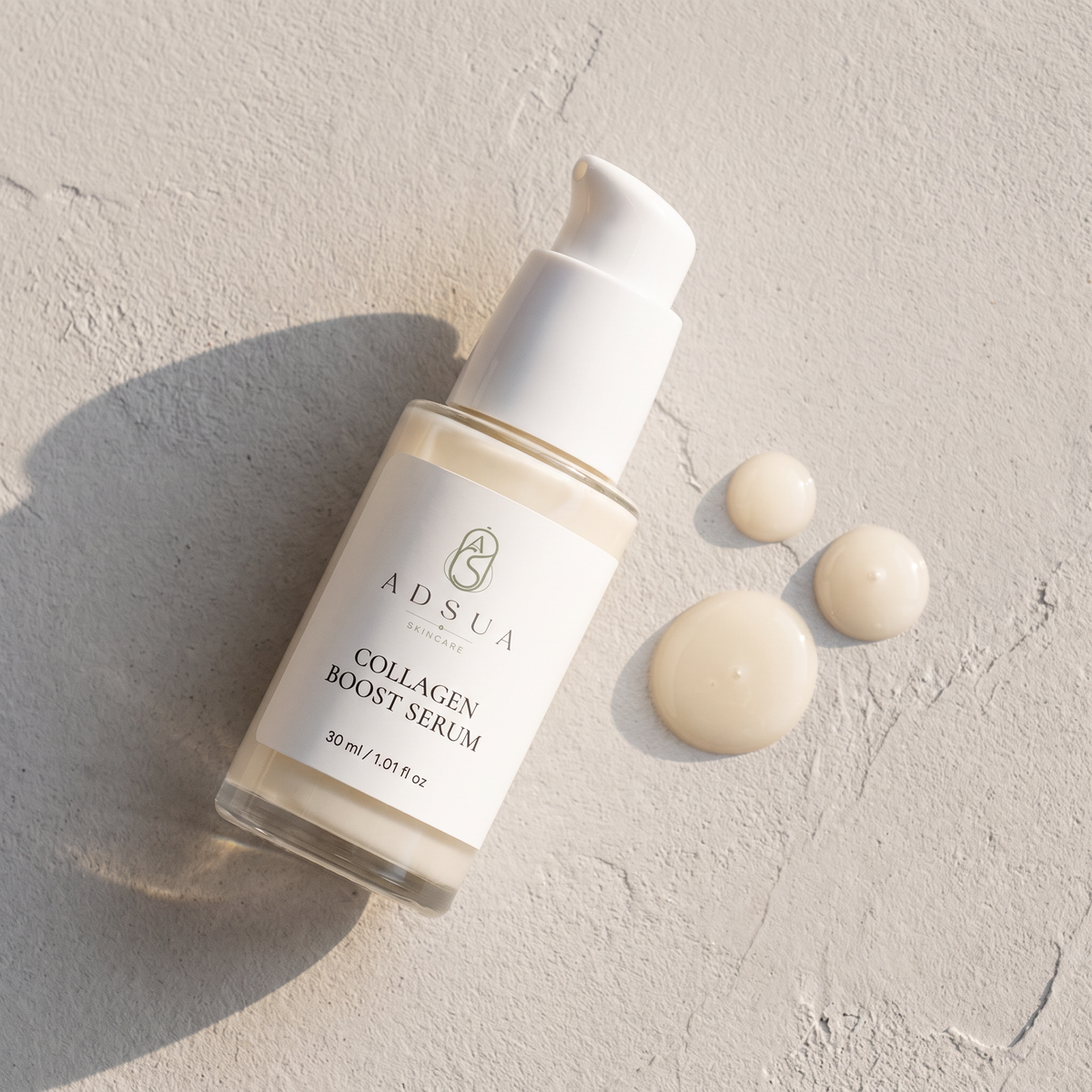 Adsua Collagen Boost Serum op lichtgrijs stucwerk met drie glanzende serumdruppels