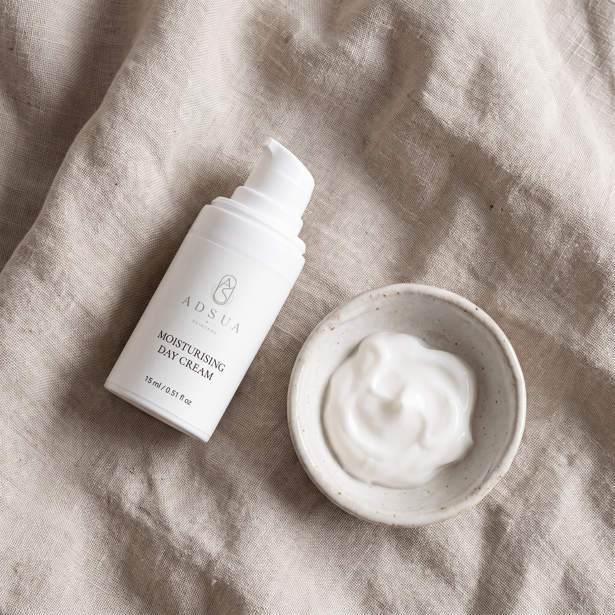 Adsua Moisturising Day Cream van 15 ml op linnen, met een schaaltje witte creme ernaast