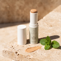 Adsua Sun Protection SPF50 Stick with tint geopend op travertijn in de zon, met een veeg van de getinte balm en een takje munt