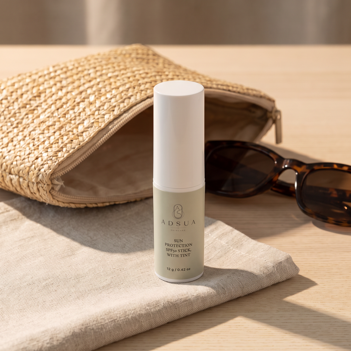 Adsua Sun Protection SPF50 Stick with tint op linnen naast een gevlochten strotasje en een zonnebril